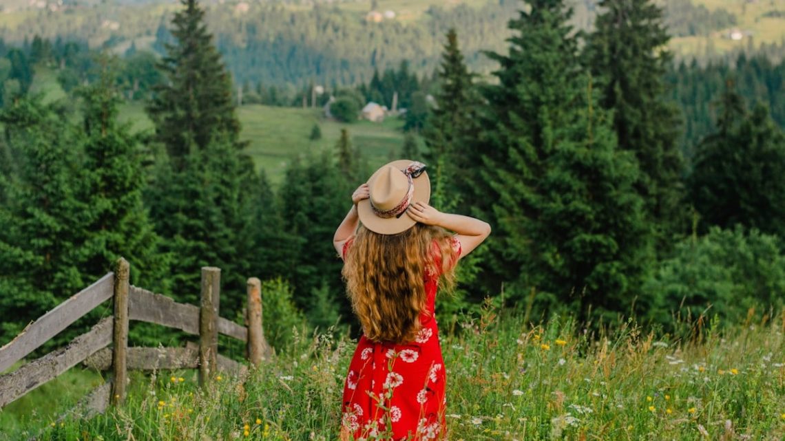 woman in red and white floral dress wearing brown sun hat standing on green grass field
