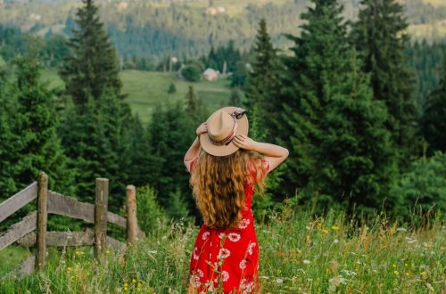 woman in red and white floral dress wearing brown sun hat standing on green grass field
